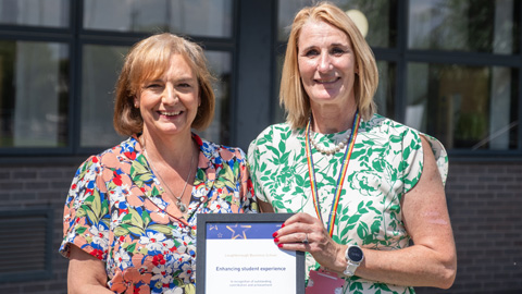 Two people in floral dresses standing outdoors in front of a building with large windows, holding a framed certificate